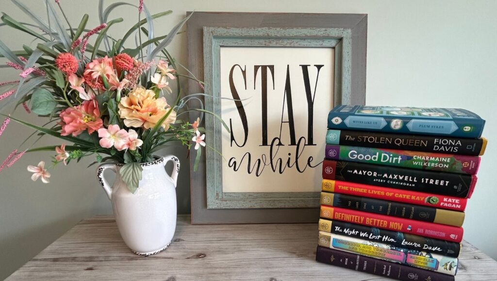 stack of colorful books and vase of flowers next to a sign that says stay a while