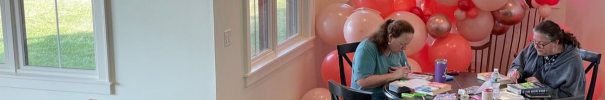 Two women bedazzling books at a Cozy Girls Getaways retreat table, surrounded by pink and rose-gold balloons.